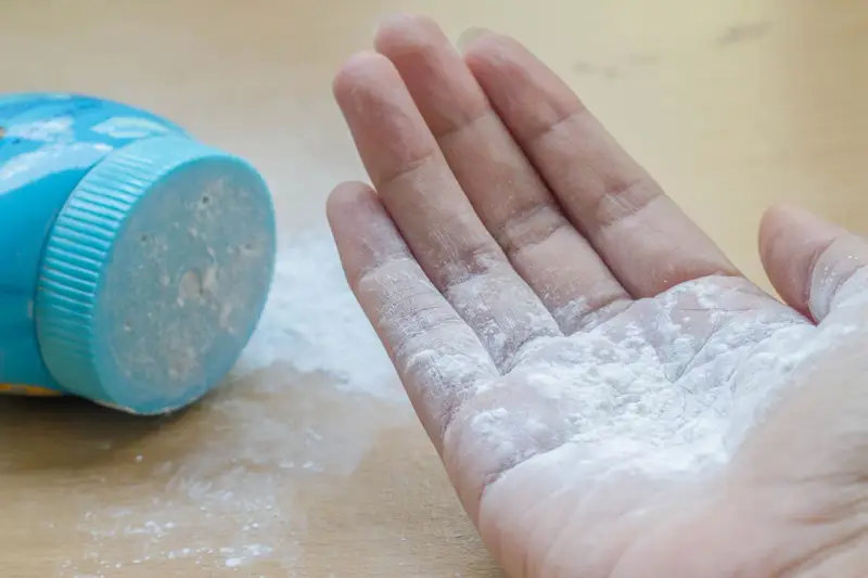 Mano con borotalco bianco sparso, vicino al contenitore blu. Attenzione: il borotalco è cancerogeno.
